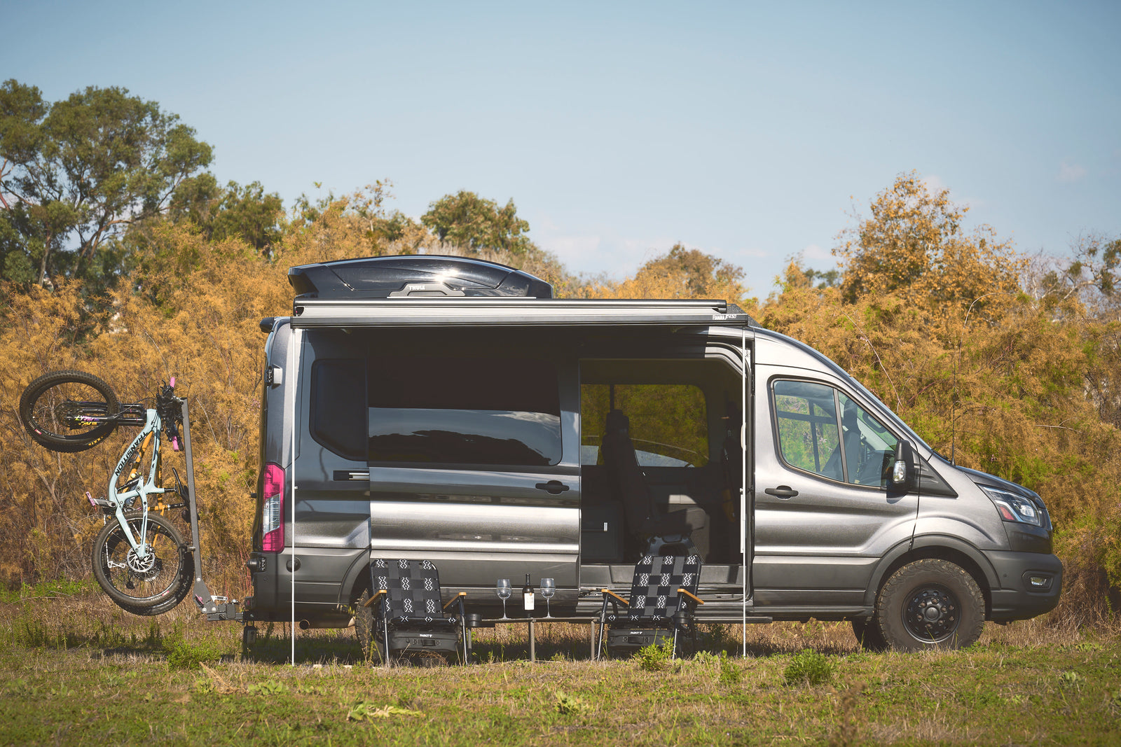 Gray RV rental camper van with a bike, extended awning, and outdoor seating setup, parked in a grassy field.