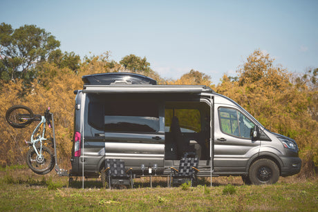 Gray RV rental camper van with a bike, extended awning, and outdoor seating setup, parked in a grassy field.