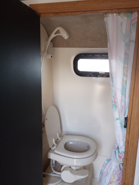 Compact RV rental bathroom: white toilet, wall-mounted shower head, small window, and floral shower curtain.