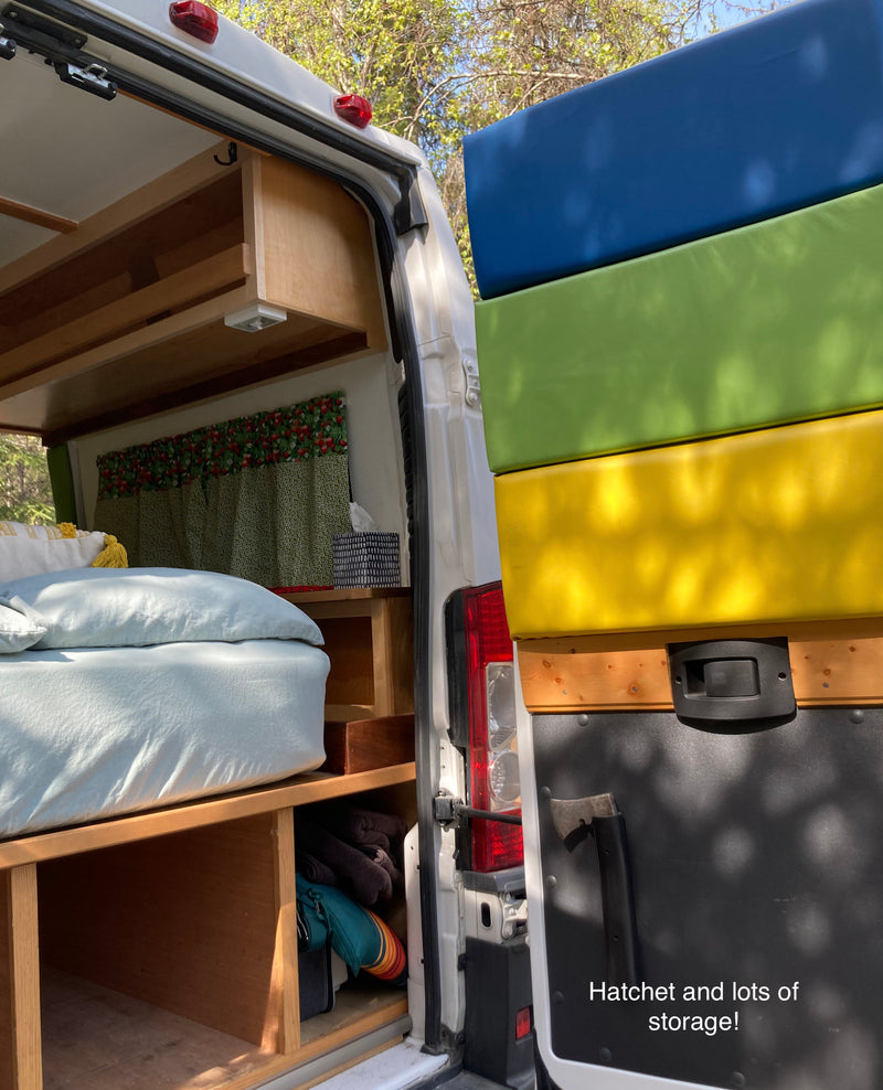 This RV rental shows a cozy camper van interior with a bed, storage shelves, and colorful exterior cushions, plus a hatchet for camping. — photo 7