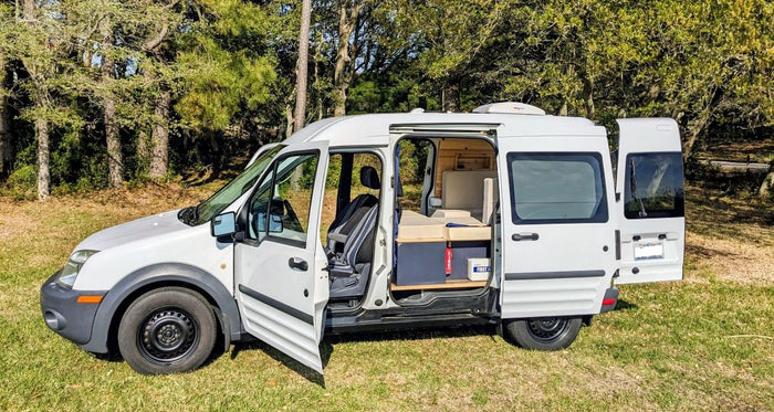 This white camper van, perfect for rv rental, has its doors open, revealing a cozy wood-paneled interior in a grassy spot.