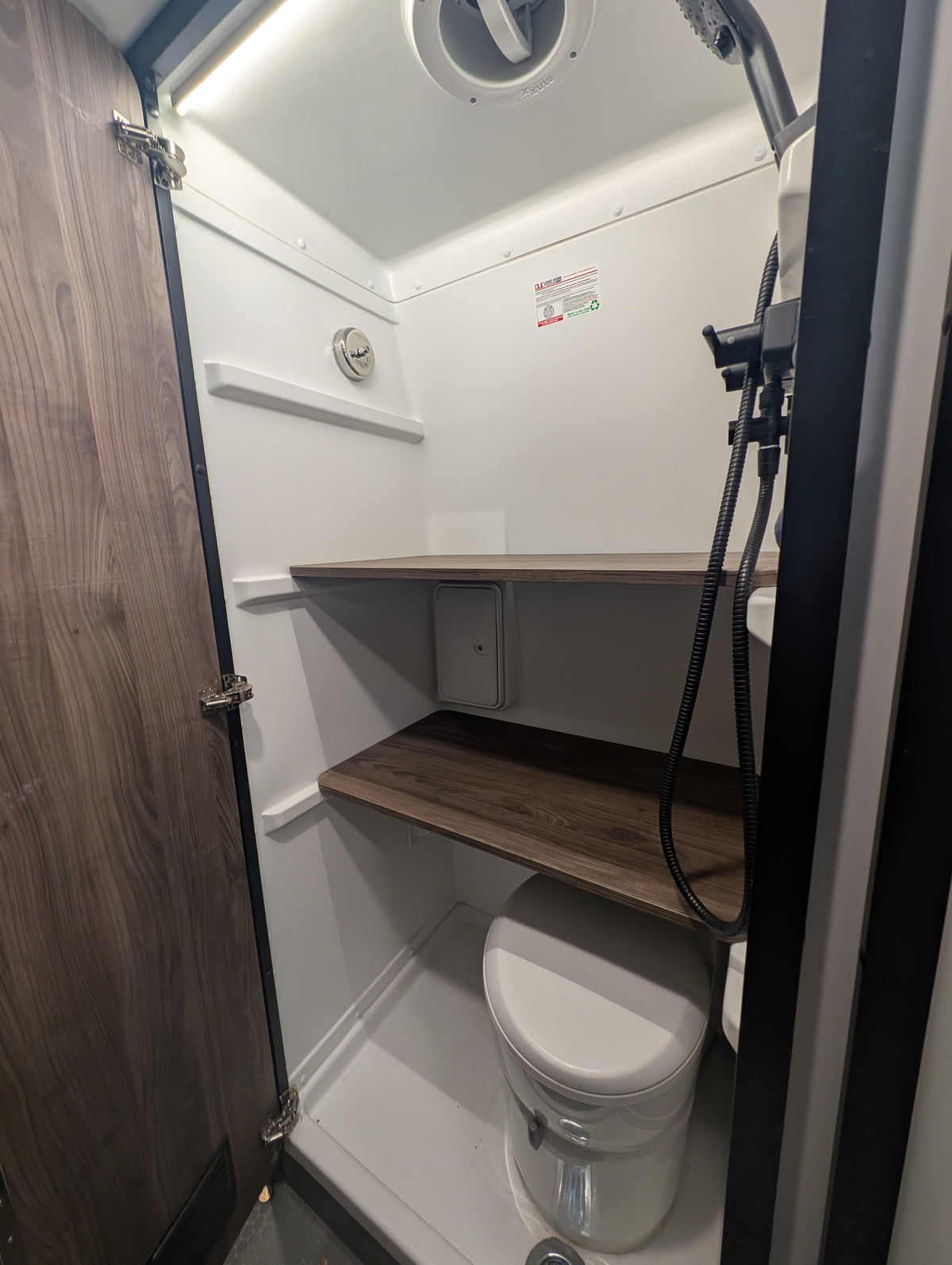 Compact RV rental wet bath. Features a toilet, shower head with hose, and two wooden shelves in a white-walled space. — photo 8