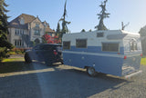 An SUV tows a classic blue and white travel trailer on a gravel driveway in front of a large house. Great for an RV rental.
