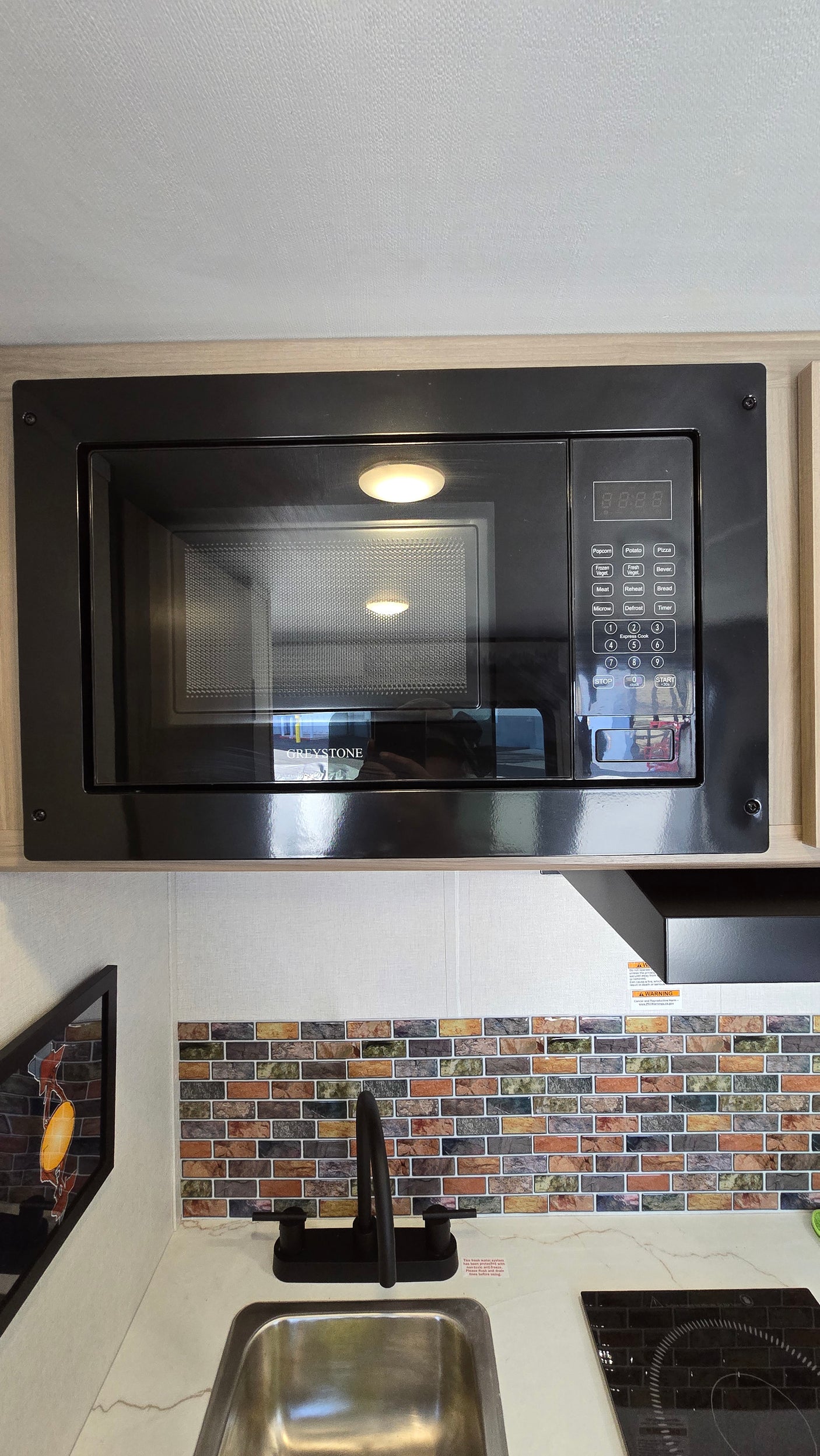 Modern rv rental kitchen featuring a black Greystone microwave, sink with faucet, induction cooktop, and colorful brick backsplash.