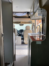 RV rental interior looking forward, showing kitchen, driver/passenger seats, and overhead storage. Vehicle parked.