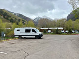 White RV rental van parked in a scenic mountain lot with snow-dusted peaks and lush trees. Ready for travel!