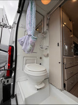 This compact rv rental bathroom has a white toilet, shower, mirror, and a tied-back curtain. Light cabinetry is on the right.