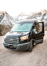 Black Ford Transit van, an RV rental, on a dirt road with snowy Utah mountains, ideal for travel and camping.