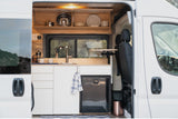 RV rental: Bright, modern camper van kitchen with sink, under-counter fridge, and wood cabinets, ready for travel.