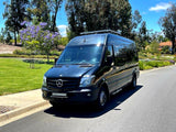 Black Mercedes Sprinter van, likely an RV rental, parked on a sunny street next to green grass and trees.