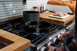 Preparing a meal in an RV rental kitchen. Hands turn stove knobs; cast iron skillet heats over blue flame, chicken prepped.