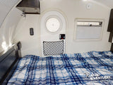 RV rental interior features a cozy bed with blue plaid comforter, round door, window, and wall-mounted controls.