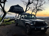 A black Jeep Gladiator with deployed RV rental rooftop tent at a coastal campsite. Ideal for adventure travel by the ocean at dusk.