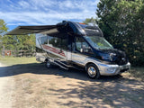 Explore nature with this Compass AWD RV rental. This black and silver motorhome is parked with its awning out near a wildlife trail.