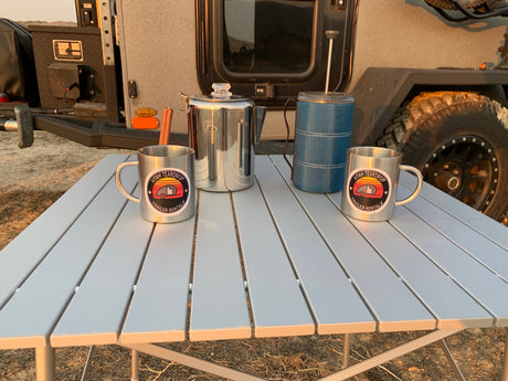 Morning coffee by your Utah Teardrop RV rental! Two custom mugs, percolator, & French press on a camp table.