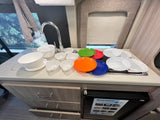 Explore an RV rental kitchen: counter, sink, cooktop, plates, bowls, and a compact fridge. Fully equipped for adventures!