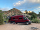 Scenic RV rental setup: red camper van with bikes, leveled for desert camping. Rocky mountains & sunset sky.