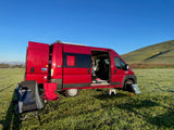 Vibrant red camper van (rv rental) with open doors, parked on a dewy grassy field beside a scenic hill. Perfect for outdoor adventures.