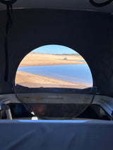 Inside an RV rental pop-top camper, a serene lake and sandy shore with a distant truck are framed by the window.