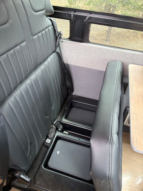 This RV rental interior features black passenger seats with seatbelts, under-seat storage bins, and a window with a nature view.
