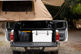 This pickup truck, perfect for an RV rental adventure, sports a tan rooftop tent, a cooler, and storage in its open bed.
