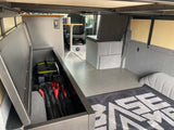RV rental interior: Open textured gray storage compartment reveals a power station & folded chairs, plus gray floor and seating.