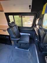 RV rental interior: A black leather captain's chair with armrests, custom wood cabinetry, and large windows in a campervan.