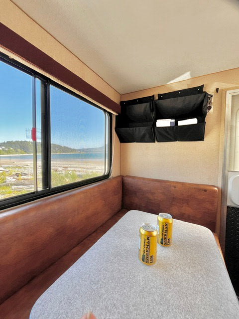 RV rental interior: cozy dining nook with a scenic water view. Features built-in bench seating, table, and wall organizers. Two cans on table.