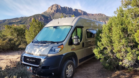 Golden Ram ProMaster RV rental van sits on a dirt path, nestled among green trees with majestic mountains in the background.