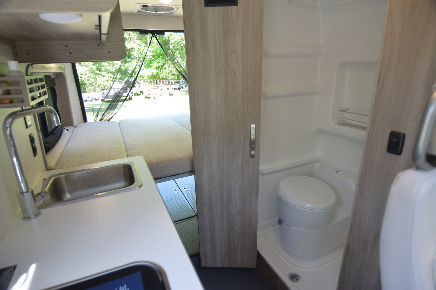 View inside an RV rental: kitchen sink on left, bed in background, and a compact bathroom with toilet and shower on the right.