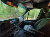 Inside an RV rental van at a forest campsite: dashboard with digital display, leather passenger seat, and outdoor table.