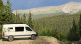 Cream RV rental camper van parked on a dirt pull-off, overlooking vast mountain ranges and dense pine forests.