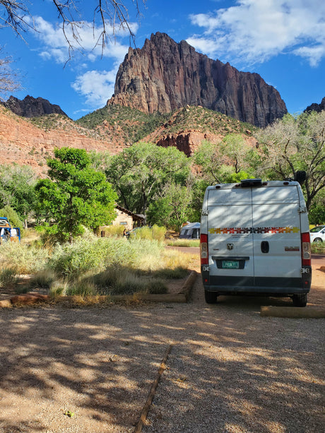 White RV rental van at a scenic national park campsite, framed by towering red rock mountains & green trees.