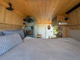 Inside a spacious RV rental: a cozy bed, warm wood paneling, and a view into the equipped living area.