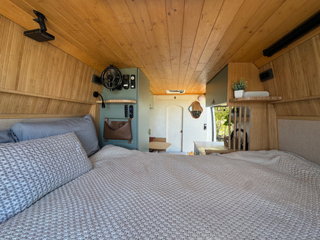 Inside a spacious RV rental: a cozy bed, warm wood paneling, and a view into the equipped living area.