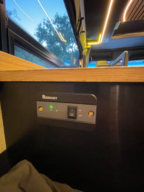 Inside an RV rental, a Renogy control panel is active with a green indicator light, alongside warm interior LED lighting.
