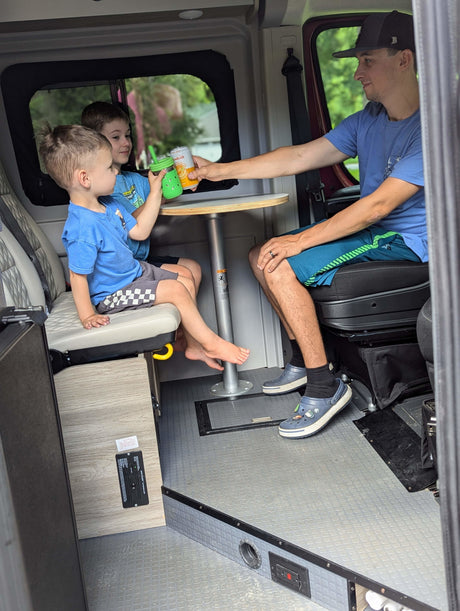 Inside a spacious rv rental, a dad gives drinks to two happy boys seated at a table.