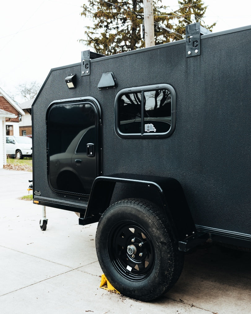 Close-up of a compact, black textured overland trailer, ideal for RV rental adventures. Features a door, window, and rugged wheel.