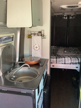 RV rental interior: Compact camper van kitchen with sink, cooktop, and cozy sleeping area beyond.