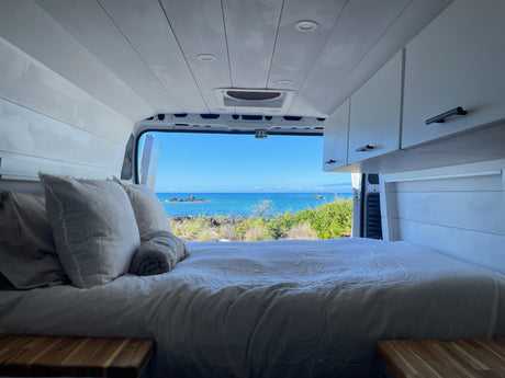 Inside an RV rental: a comfy bed overlooks a stunning blue ocean, rocky shore, and green plants. Ideal for van life travelers.