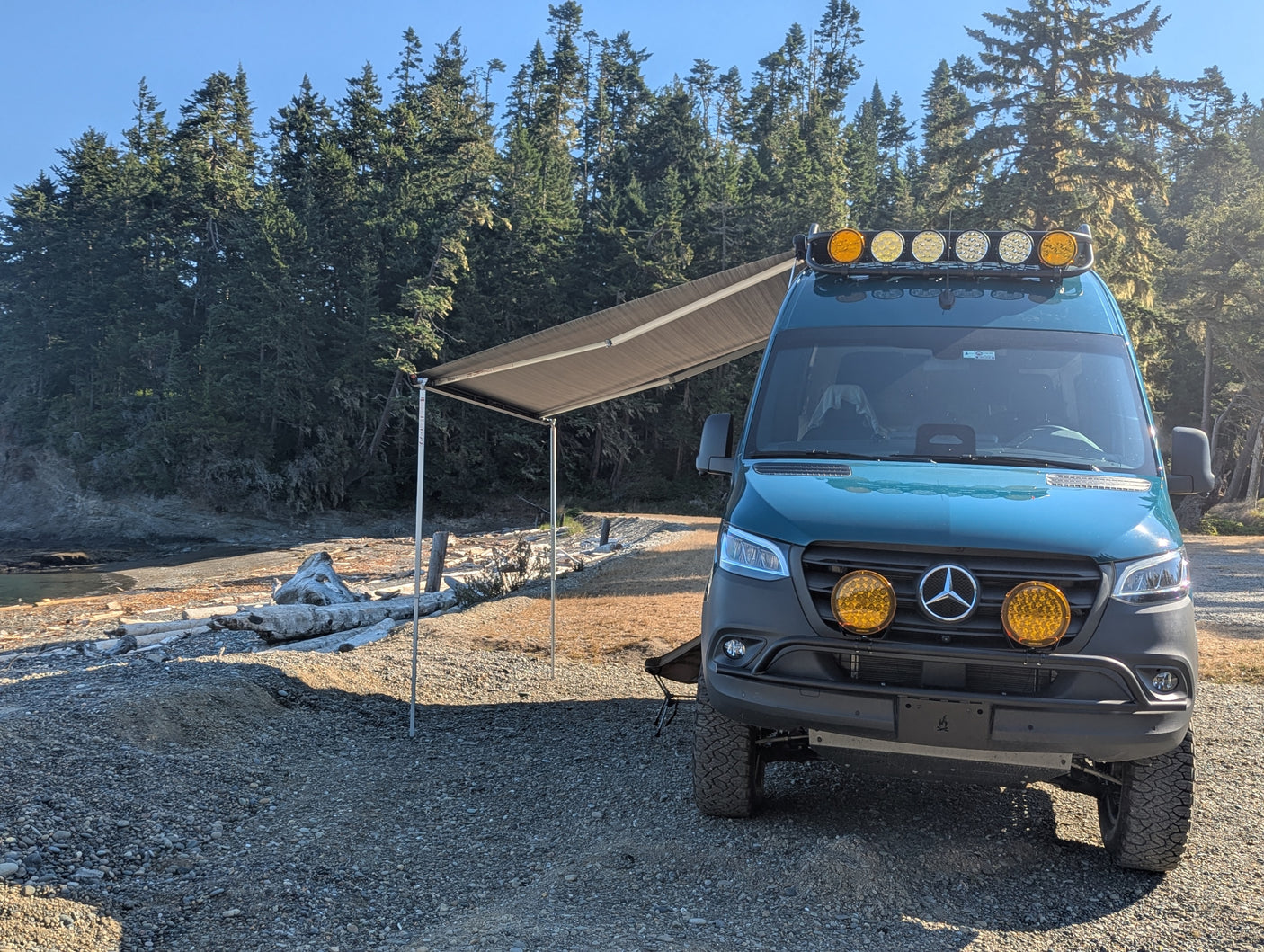 Off-road teal Mercedes Sprinter rv rental with awning extended, parked on a sunlit gravel beach by a dense forest.