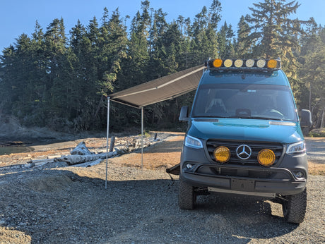 Off-road teal Mercedes Sprinter rv rental with awning extended, parked on a sunlit gravel beach by a dense forest.