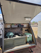 Open RV rental hatch reveals a compact outdoor kitchen: sink, portable fridge, storage, speakers, and paper towels.
