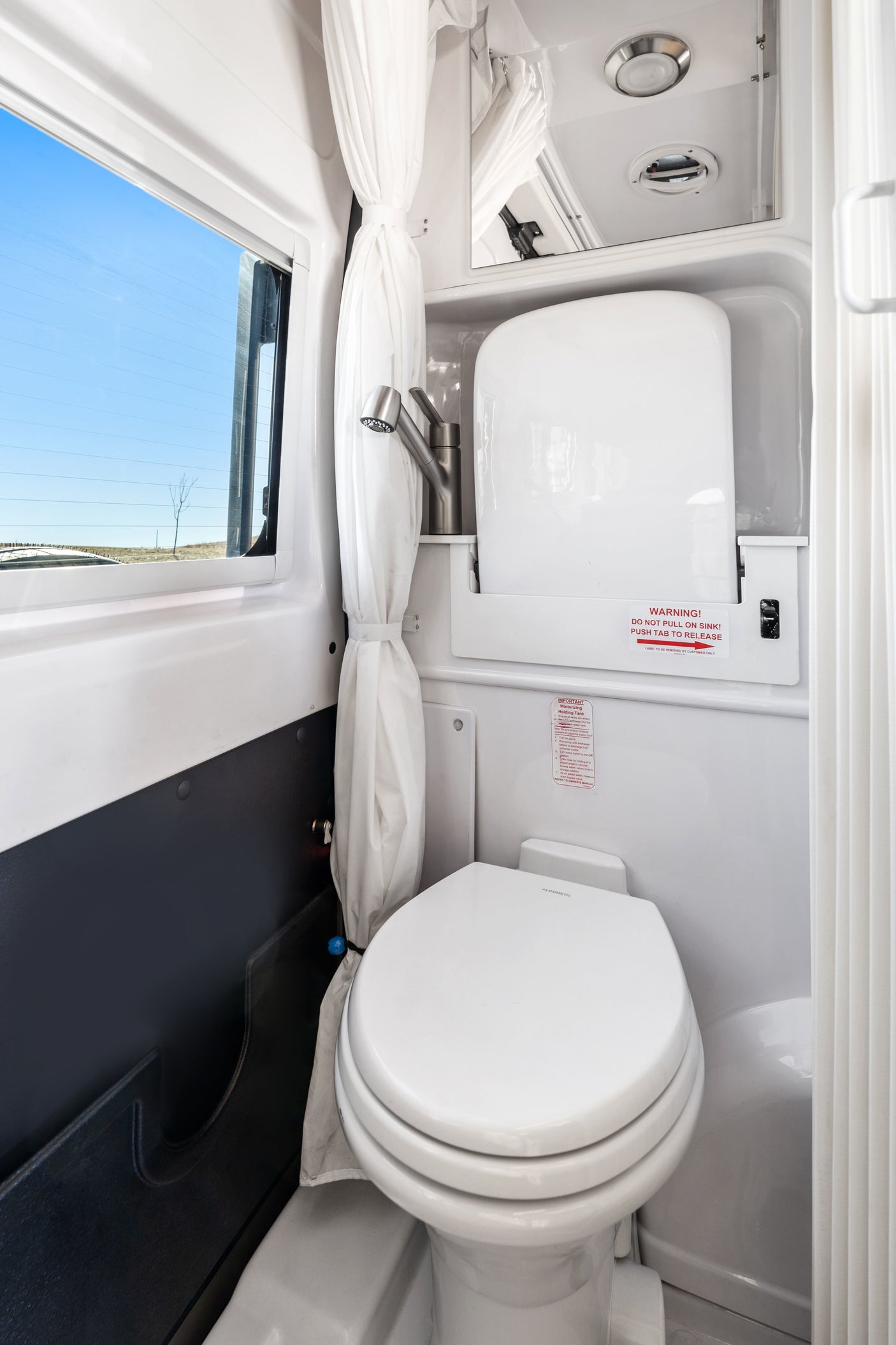 Compact RV rental wet bath with Dometic toilet, pull-out sink, and shower curtain, offering a clear view out the window.