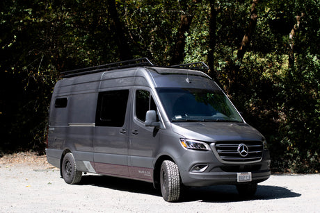 A gray Mercedes Sprinter camper van for RV rental, ready for outdoor adventure on a gravel road by lush trees.