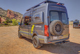 Adventure-ready gray Sprinter RV rental with off-road tires and roof rack at a scenic red rock park.
