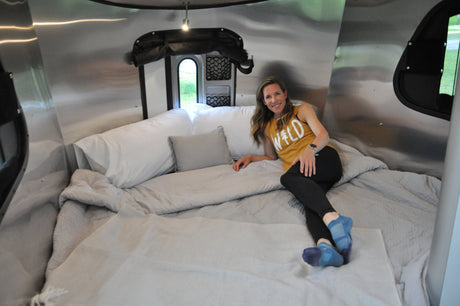 Woman smiles lounging in a cozy RV rental bed, with sleek metallic interior and outdoor views. Perfect for travel.