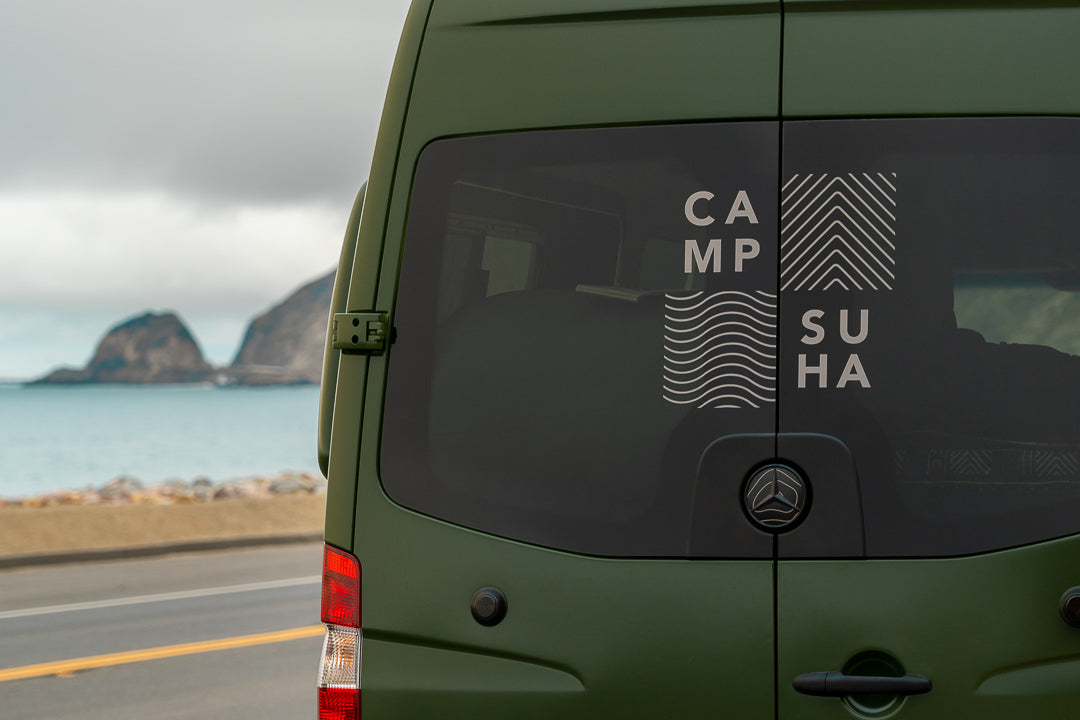 Green camper van rv rental displays 'CAMPSUHA' on back window, parked by the ocean with rocks in distance.