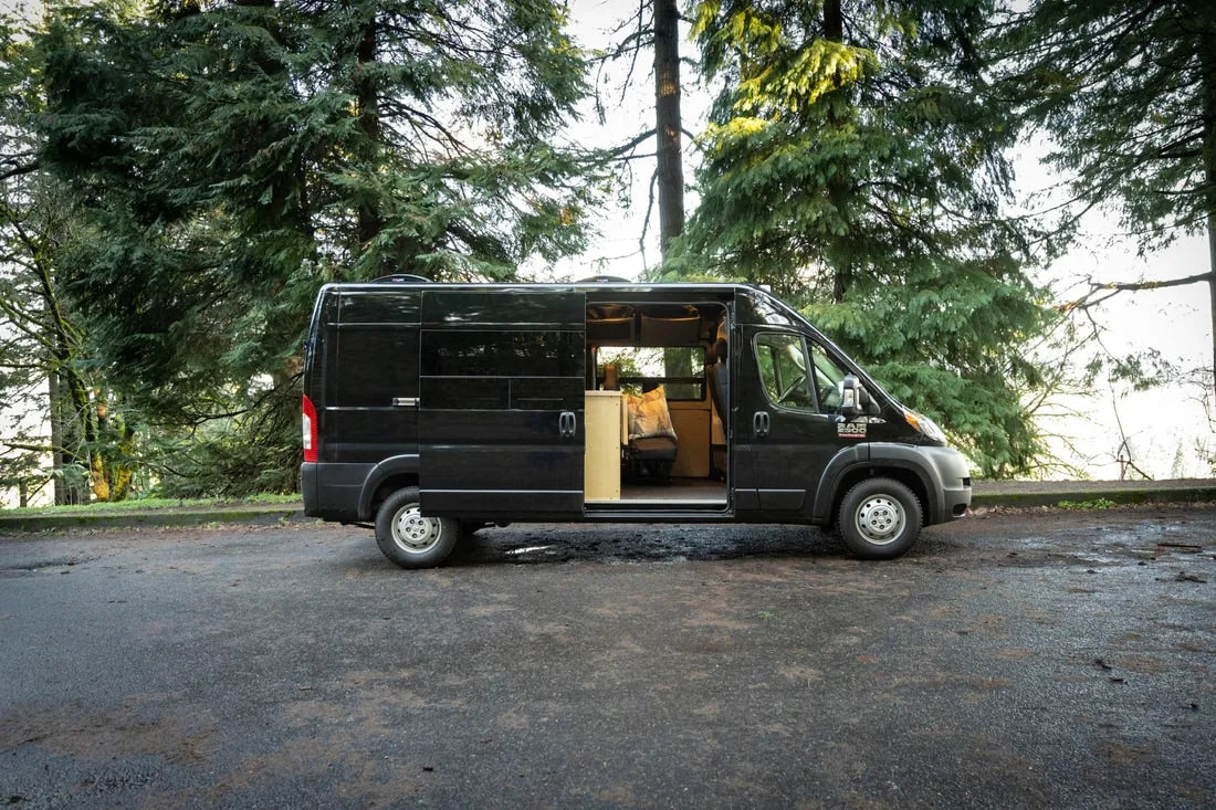 Black Ram ProMaster camper van RV rental parked among tall pine trees with side door open, showing the converted interior.