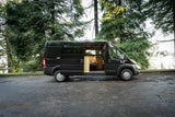 Black Ram ProMaster camper van RV rental parked among tall pine trees with side door open, showing the converted interior.